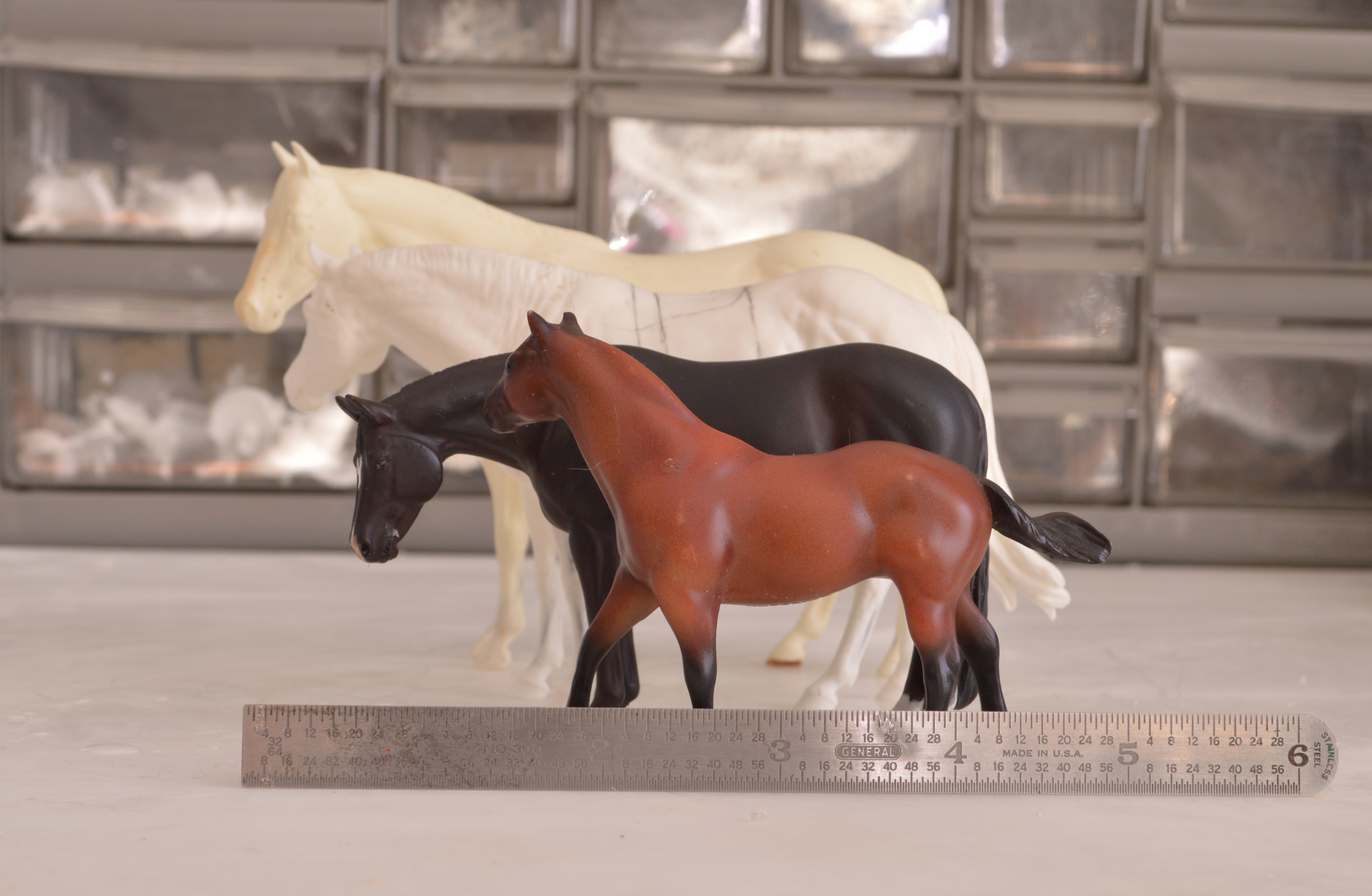 Four model horses viewed from left side, smallest in front to largest in back: bay breyer G1 quarter horse, black curio stock horse, white resin Chino, white resin miss mattie. Six inch metal ruler for scale