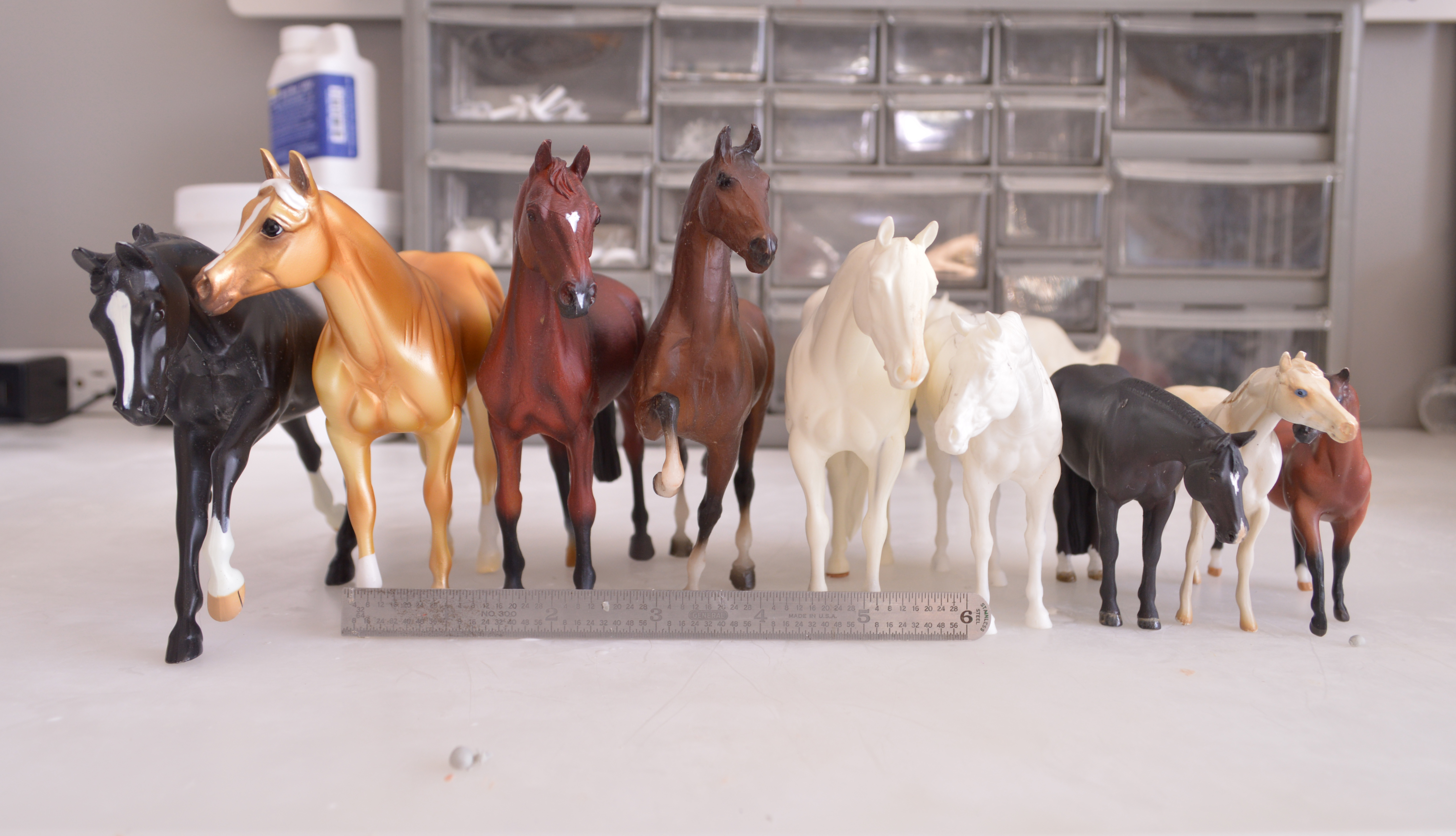 Line of nine model horses viewed from the front and arranged left to right, tallest to shortest: black pebble loping stock horse, palomino standing stock horse by breyer, chestnut Collecta warmblood, brown little bits morgan, yellow-ish unpainted resin stock horse, white unpainted stock horse significantly smaller than previous models, much smaller black stock horse, cream colored stone chip, bay breyer G1 quarter horse. Six inch silver metal ruler for scale