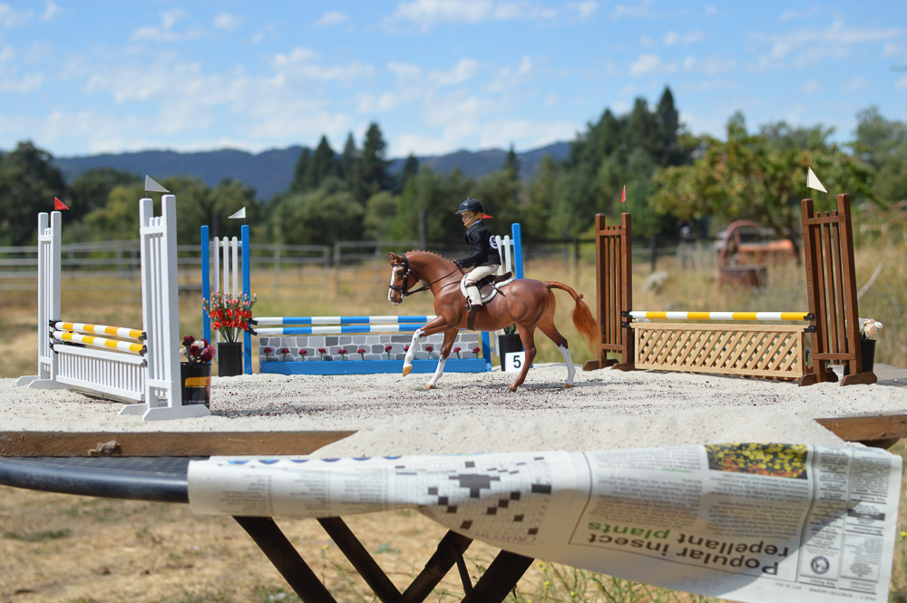 Chestnut pony cantering towards a jump on a table with the edges of the miniature scene visible.