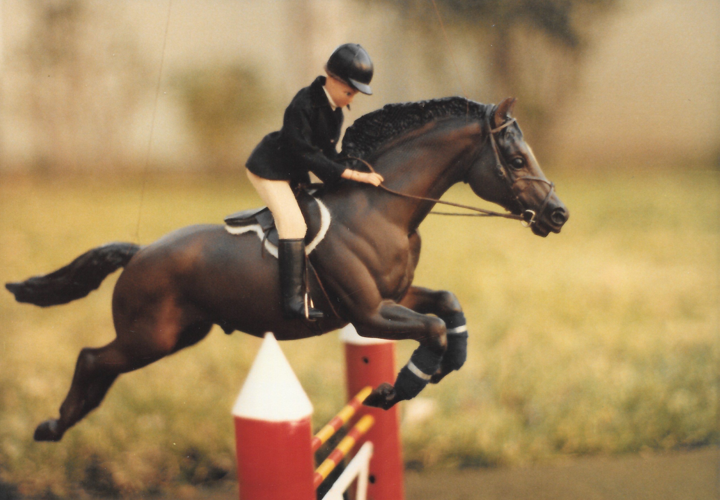 Breyer Jumper in dark bay set up for show jumping with a doll wearing a black jacket and looking down. The horse is suspended by a thread, facing right.